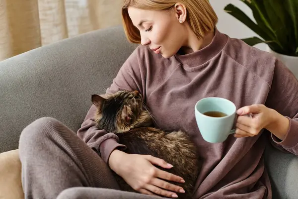 Una donna chic con i capelli corti si rilassa su un divano, assaporando il caffè e coccolando il suo gatto. — Foto stock