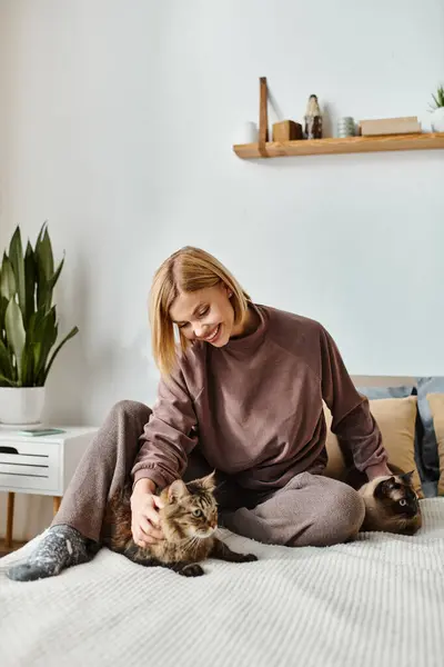 A woman with short hair sits on a bed, petting her cats. — Stock Photo