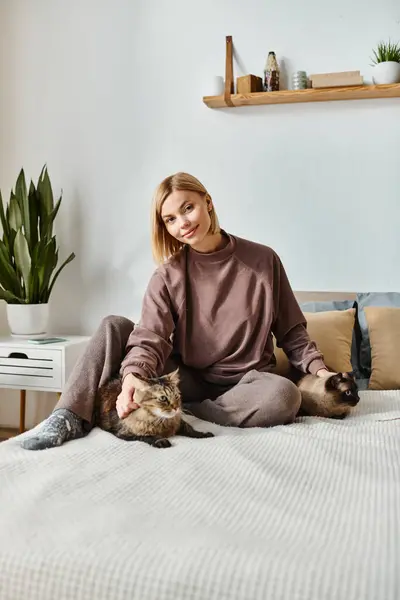 Una mujer con el pelo corto serenamente sentado en una cama, acariciando suavemente a un gato calico a su lado. - foto de stock