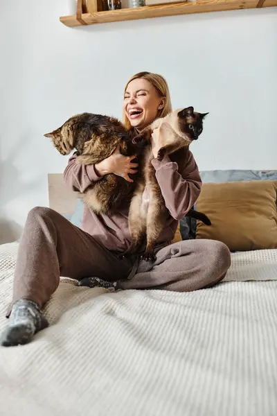 Жінка з коротким волоссям мирно сидить на ліжку, тримаючи двох котів поруч з нею. — стокове фото