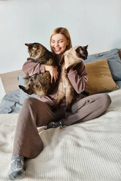 Жінка з коротким волоссям сидить на ліжку, тримаючи двох котів поруч з нею, насолоджуючись мирною миттю вдома. — стокове фото