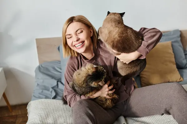 Une femme aux cheveux courts assise sur un lit, tenant tendrement deux chats dans ses bras, profitant d'un moment paisible à la maison. — Photo de stock