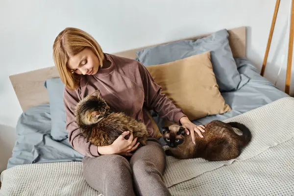 Uma mulher com cabelo curto pacificamente sentada em uma cama, segurando dois gatos em seus braços, desfrutando de momentos tranquilos em casa. — Fotografia de Stock