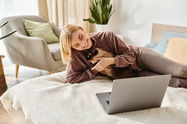 Una donna elegante con i capelli corti si rilassa su un letto, assorto nel suo computer portatile mentre il suo gatto coccola accanto a lei. — Foto stock