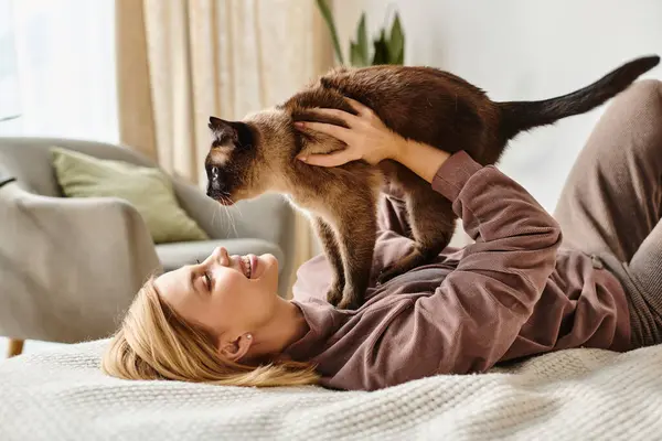 Una mujer con el pelo corto se relaja en una cama, acunando a su gato en sus brazos, exudando paz y satisfacción. - foto de stock