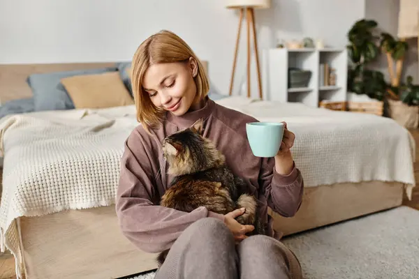 Una mujer atractiva con el pelo corto se sienta en el suelo, sosteniendo una taza mientras se acaricia con su gato. - foto de stock