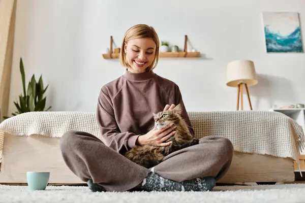 Una mujer con el pelo corto sentada en una cama, acunando a un gato en sus brazos en un momento pacífico y cariñoso. - foto de stock