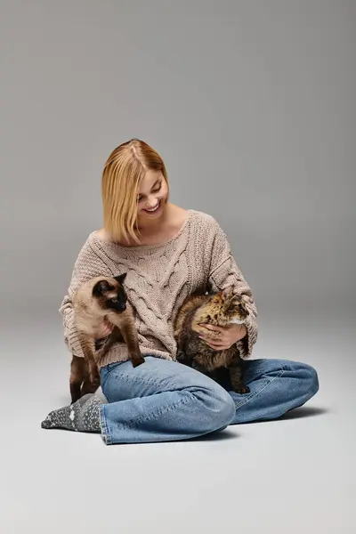 Una donna con i capelli corti siede sul pavimento, teneramente stringendo due gatti tra le braccia, mostrando un momento sereno e pacifico.. — Foto stock