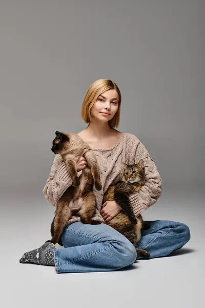 Una donna dai capelli corti si siede sul pavimento, cullando due gatti tra le braccia, incarnando un momento di pace e contenuti a casa.. — Foto stock