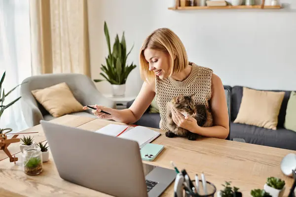 Una mujer elegante con el pelo corto trabaja en su computadora portátil en una mesa mientras su compañero felino peludo se sienta a su lado. - foto de stock