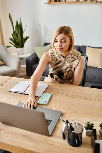 Una mujer con el pelo corto sentado en una mesa, absorto en el uso de una computadora portátil mientras su gato se sienta cerca. - foto de stock