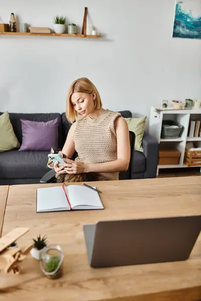 Eine Frau mit kurzen Haaren sitzt an einem Tisch, in ihr Handy vertieft, während ihre Katze in der Nähe faulenzt.. — Stockfoto