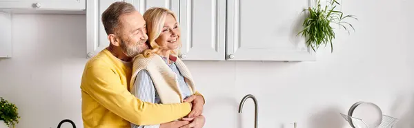 A mature man and woman in cozy homewear share a tender hug in a warm kitchen setting. — Stock Photo