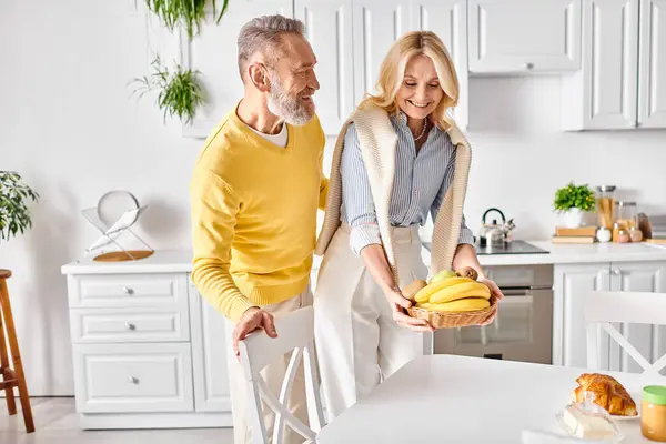 Un hombre y una mujer maduros están en su acogedora cocina, sosteniendo plátanos y compartiendo un momento dulce juntos. - foto de stock