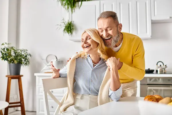 A mature loving couple, dressed in cozy homewear, share a moment of genuine laughter and joy in their kitchen at home. — Stock Photo