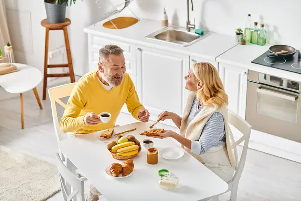 A mature man and woman enjoy a cozy meal together at a table in their kitchen, sharing love and laughter. — Stock Photo