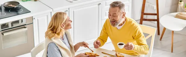 A mature loving couple enjoying a meal together at a table in cozy homewear, savoring delicious food in a warm atmosphere. — Stock Photo