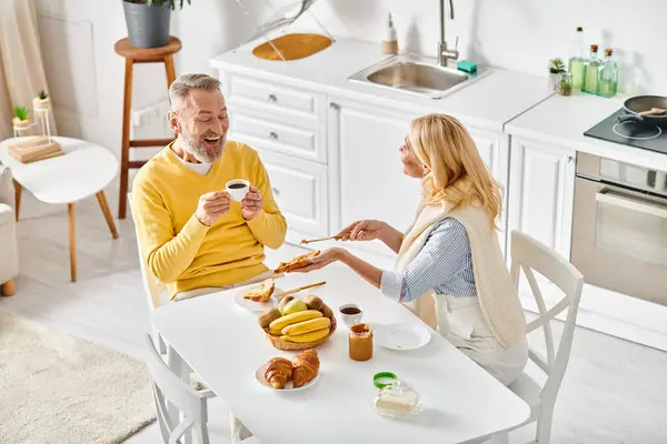 A mature loving couple in cozy homewear sitting together at a kitchen table, enjoying a moment of togetherness at home. — Stock Photo