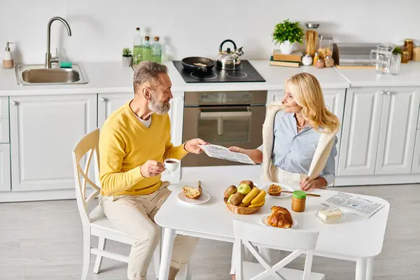 Un uomo e una donna maturi in accogliente biancheria da casa seduti insieme a un tavolo da cucina, godendo di un momento di tranquillità insieme. — Foto stock