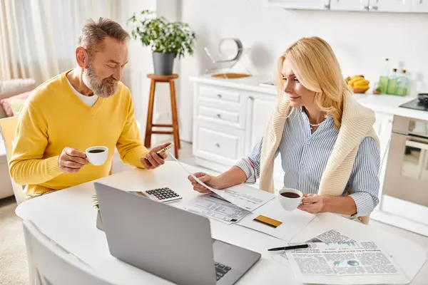 A mature loving couple in cozy homewear working together on a laptop at a table in their home kitchen. — Stock Photo