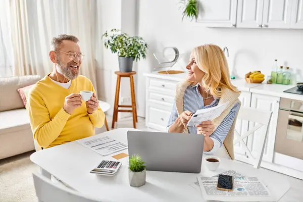 A mature loving couple dressed in homewear working together on a laptop at a table in a cozy kitchen setting. — Stock Photo