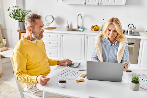 A mature loving couple in cozy homewear sitting together at a table, working on a laptop. — Stock Photo