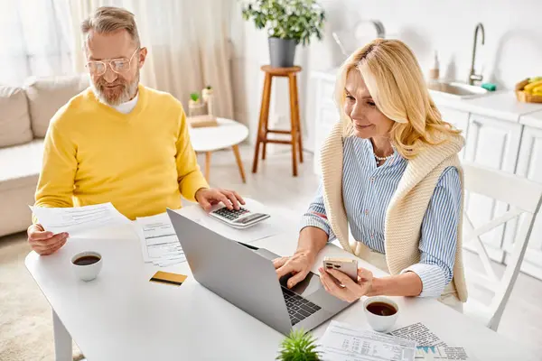 A mature man and woman in cozy homewear sit at a table, engrossed in their laptop screen, in a warm domestic setting. — Stock Photo