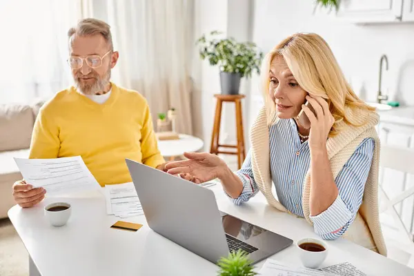 A mature loving couple in cozy homewear sitting at a table with papers and a laptop, engrossed in a productive work session. — Stock Photo