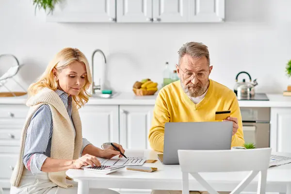 A mature loving couple in cozy homewear sitting at a kitchen table, working together on a laptop. — Stock Photo