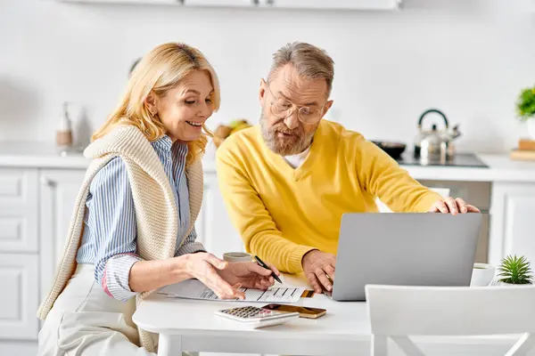 A mature loving couple in homewear sits at a table, engrossed in a laptop screen, sharing a cozy moment in their kitchen at home. — Stock Photo