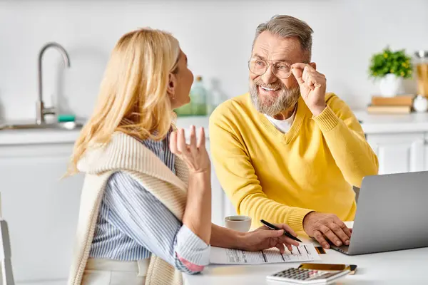 A mature loving couple in cozy homewear sitting at a table, focused on their laptop in a relaxed kitchen setting. — Stock Photo
