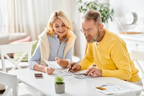 A mature loving couple in cozy homewear sit together at a table, enjoying a quiet moment together in their kitchen at home. — Stock Photo