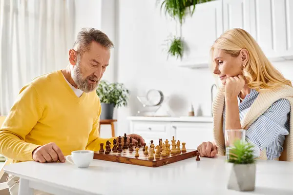 Una pareja madura y cariñosa en una acogedora ropa de casa enfocada en un juego de ajedrez, mostrando pensamiento estratégico y compromiso mutuo. - foto de stock