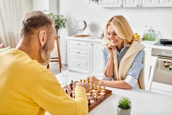 Una pareja madura y cariñosa en acogedora ropa de casa participando en un juego estratégico de ajedrez en casa. - foto de stock
