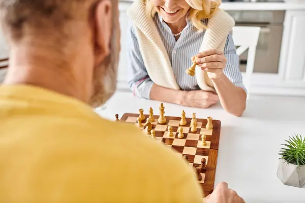 Una pareja madura y cariñosa en una acogedora ropa de casa se sienta una frente a la otra, profundamente centrada, mientras hacen estrategias y juegan un juego de ajedrez en casa.. - foto de stock