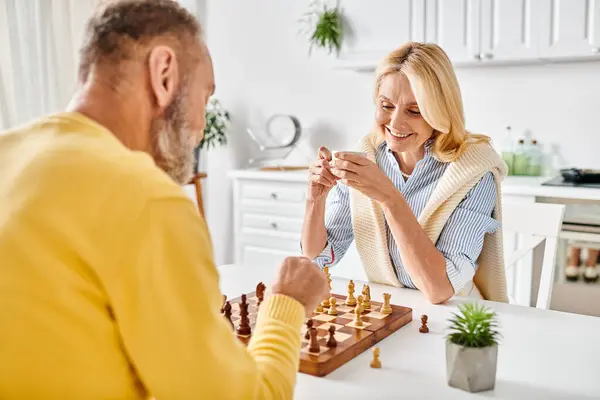 Un couple d'âge mûr dans des vêtements de maison confortables engagés dans un jeu d'échecs, se concentrant intensément sur l'échiquier alors qu'ils élaborent leur prochaine stratégie. — Photo de stock
