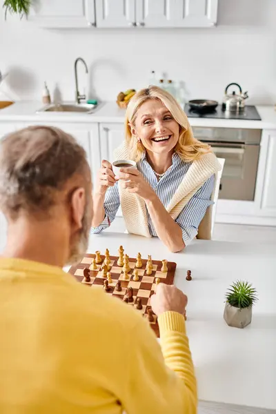 Una pareja madura y cariñosa en ropa de casa acogedora participar en un desafiante juego de ajedrez en su cocina, centrado y reflexivo. - foto de stock