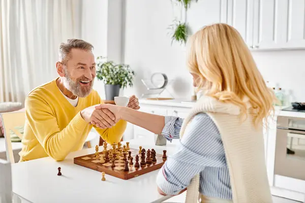 Un hombre y una mujer maduros comprometidos en un juego estratégico de ajedrez en su acogedora cocina, disfrutando de un momento de desafío intelectual y conexión. - foto de stock