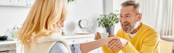A mature man and woman in cozy homewear sit at the kitchen table, shaking hands in a gesture of agreement. — Stock Photo