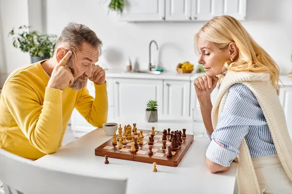 Um casal amável maduro em casa aconchegante envolvido em uma batalha estratégica de xadrez em uma mesa em sua cozinha em casa. — Fotografia de Stock