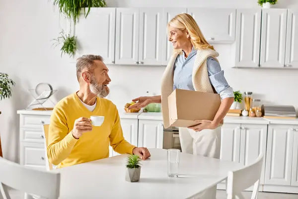 Un hombre en ropa interior acogedora sostiene una caja, mientras que una mujer con una pizza, creando un ambiente cálido en su cocina. — Stock Photo