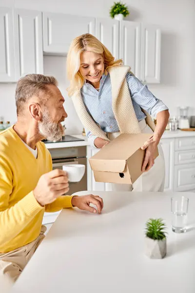 Un hombre y una mujer maduros en ropa interior acogedora están abriendo una caja en un mostrador de cocina, curioso y emocionado por su contenido. - foto de stock