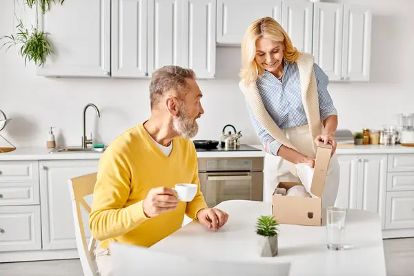 Un couple aimant mature dans des vêtements confortables ouvre avec enthousiasme une boîte de nourriture ensemble dans leur cuisine à la maison. — Photo de stock