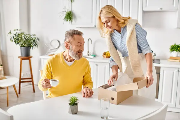 Una coppia matura e amorevole in abiti da casa accoglienti, aprendo una scatola per rivelare una pianta all'interno, condividendo un momento di gioia e scoperta nella loro cucina.. — Foto stock