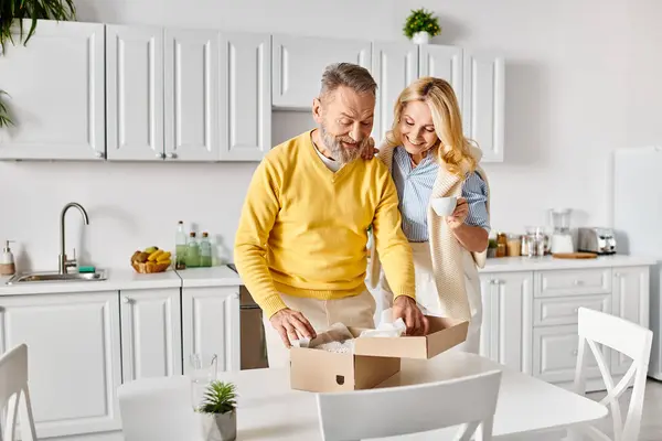 A mature man and woman in comfortable clothing open a box in a cozy kitchen, sharing a moment of curiosity and anticipation. — Stock Photo