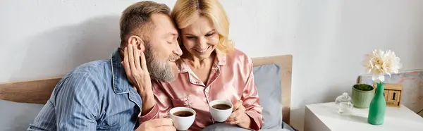A mature loving couple, dressed in cozy homewear, sit on a bed holding cups of coffee, spending quality time together. — Stock Photo