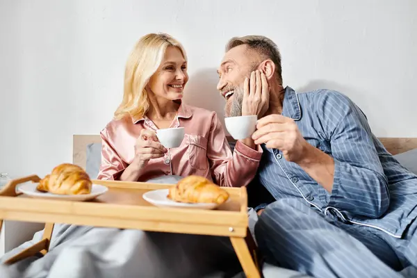 Una pareja amorosa madura en ropa de casa acogedora sentada en una cama, saboreando el café juntos en un entorno tranquilo dormitorio. - foto de stock