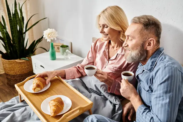 A mature couple in cozy homewear sitting on a couch, sipping coffee and indulging in pastries together in a warm and inviting atmosphere. — Stock Photo