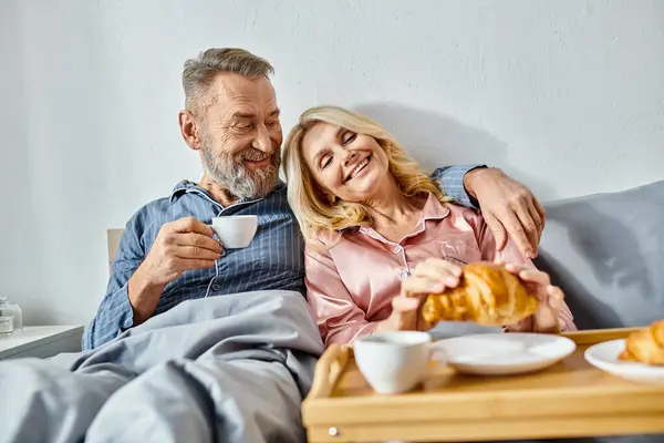 Couple d'âge mûr dans des vêtements confortables se détendre sur le canapé, partager un plateau de nourriture délicieuse. — Photo de stock