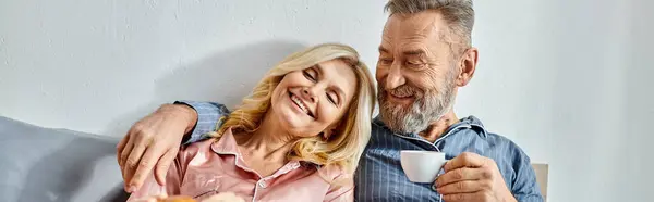 A mature man and woman in comfortable homewear sitting closely together on a cozy couch in their bedroom. — Stock Photo